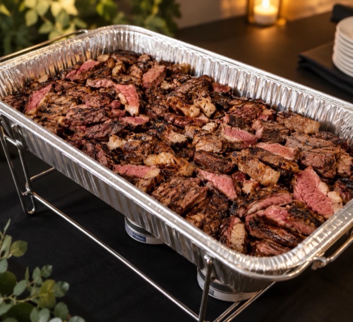 Disposable tray of sliced picanha on a chafing rack, set up buffet style for a drop-off order